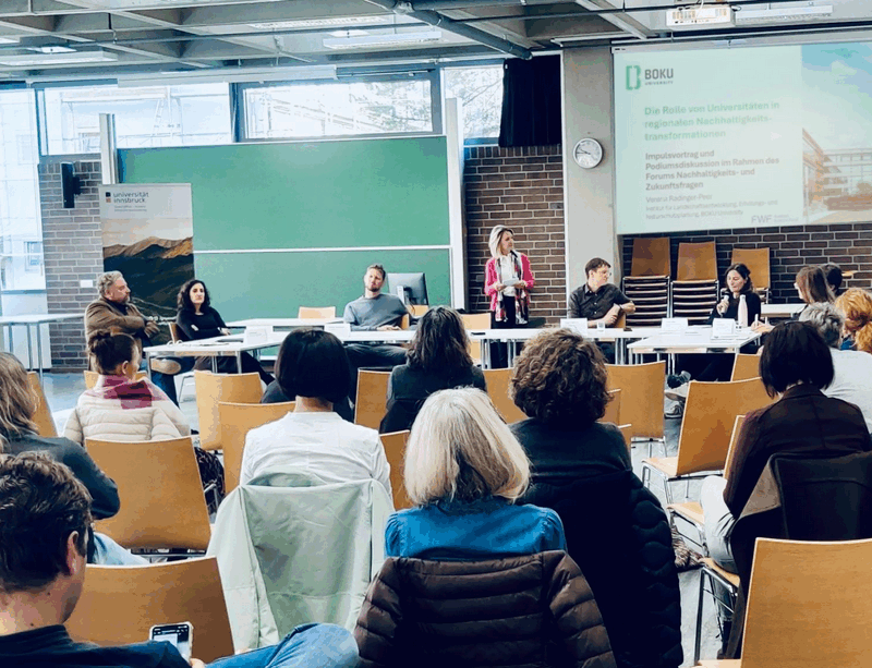 Dieses Bild zeigt eine Vortragende sowie mehrere Menschen, die am Podium sitzen. Auch ein Teil des Publikums ist zu sehen.