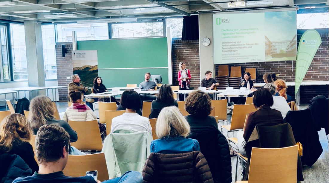 Dieses Bild zeigt eine Vortragende sowie mehrere Menschen, die am Podium sitzen. Auch ein Teil des Publikums ist zu sehen.